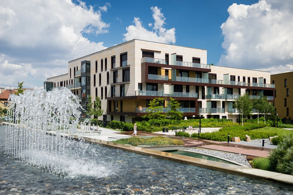 Complexe résidentiel moderne avec façades en béton et panneaux colorés, balcons avec garde-corps métalliques, espaces verts aménagés et fontaine d'eau