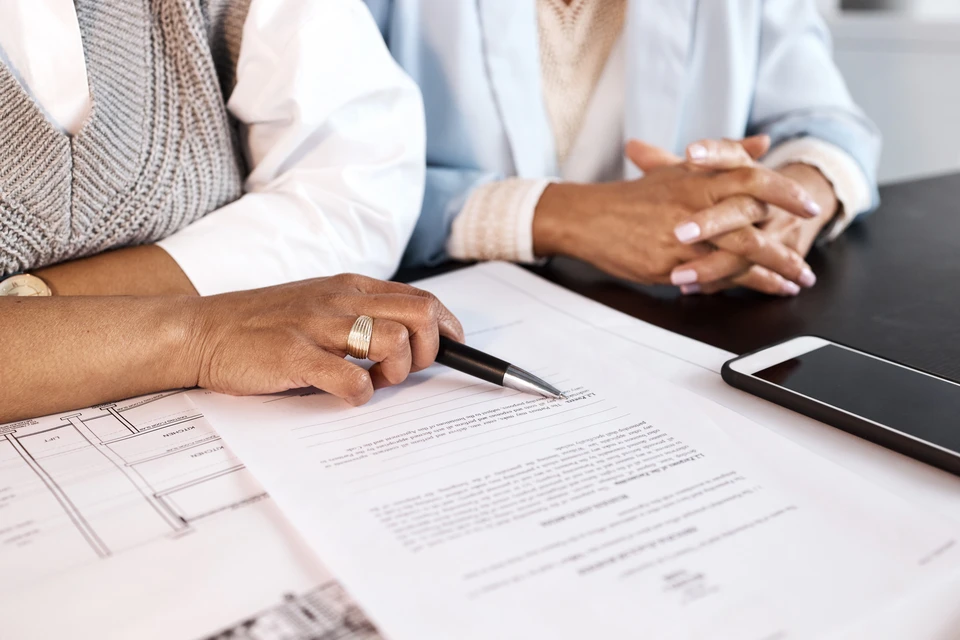 Scène d'intérieur professionnel avec deux personnes assises à une table, l'une signant des documents contractuels avec un stylo, l'autre observant les mains jointes